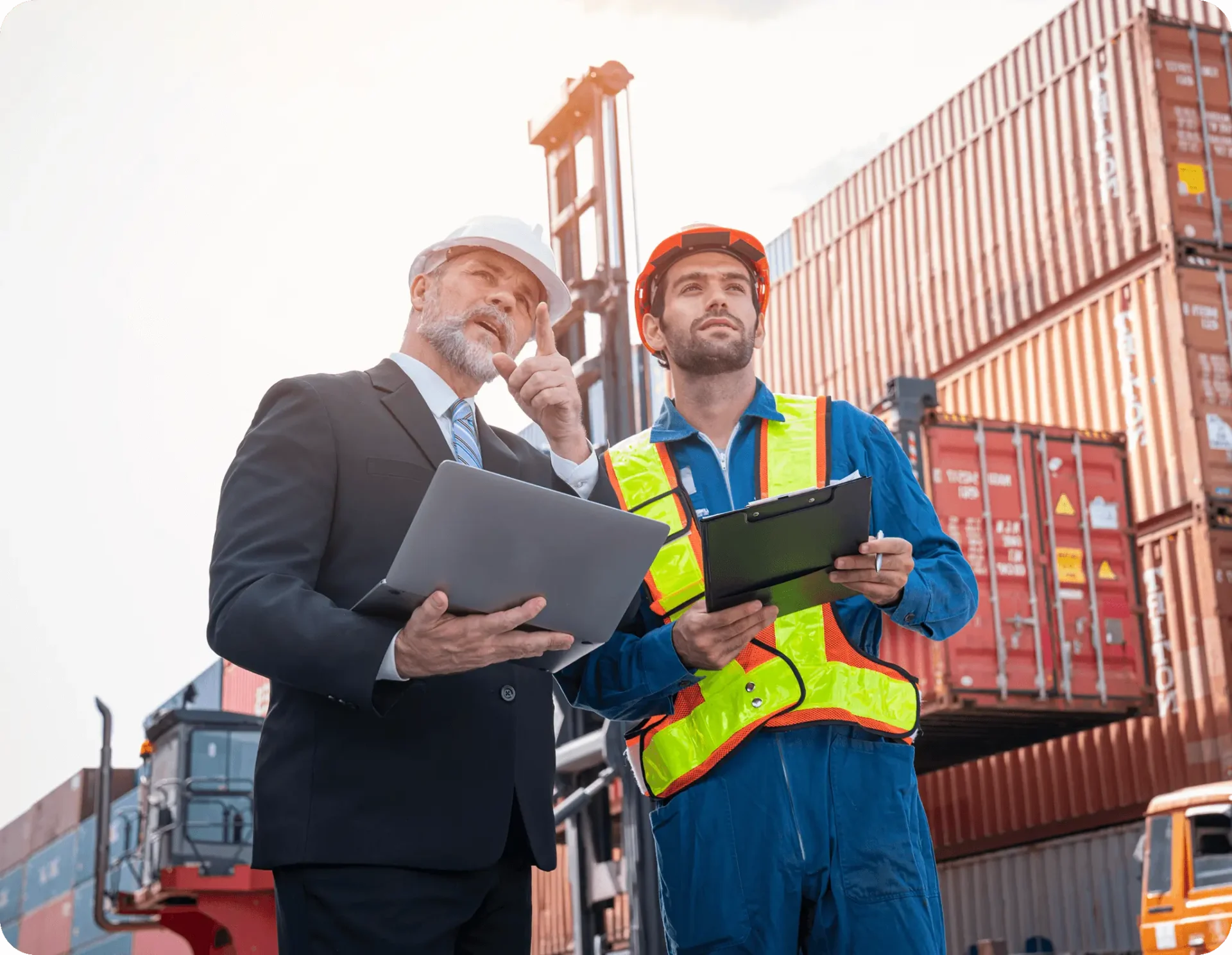 Two men at a bustling shipping port, one in a suit with a laptop from a trucking company, the other in workwear clutching a clipboard, stand before towering stacks of cargo containers.